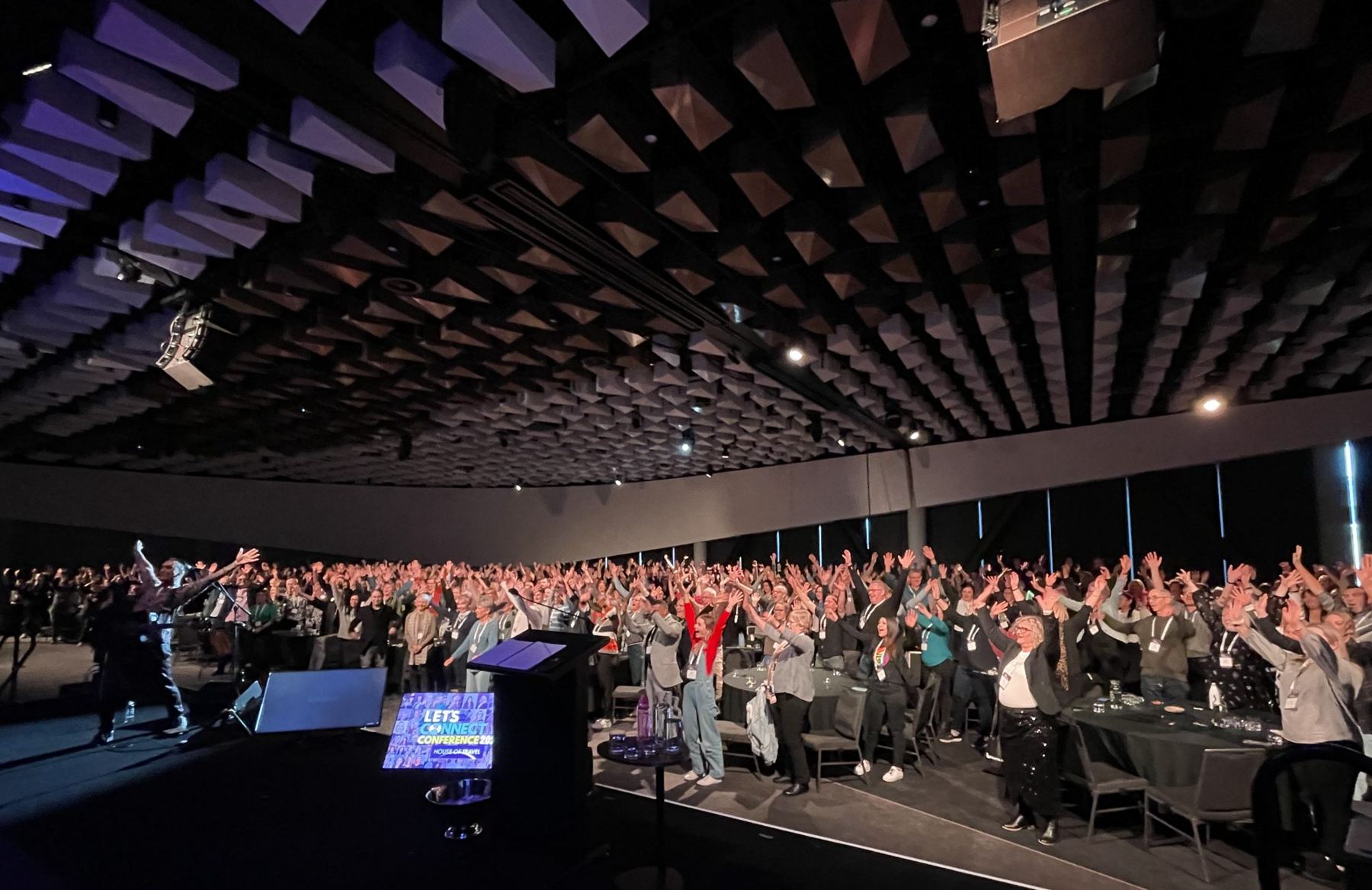 A full conference room on its feet with arms raised, led by Joel from the stage
